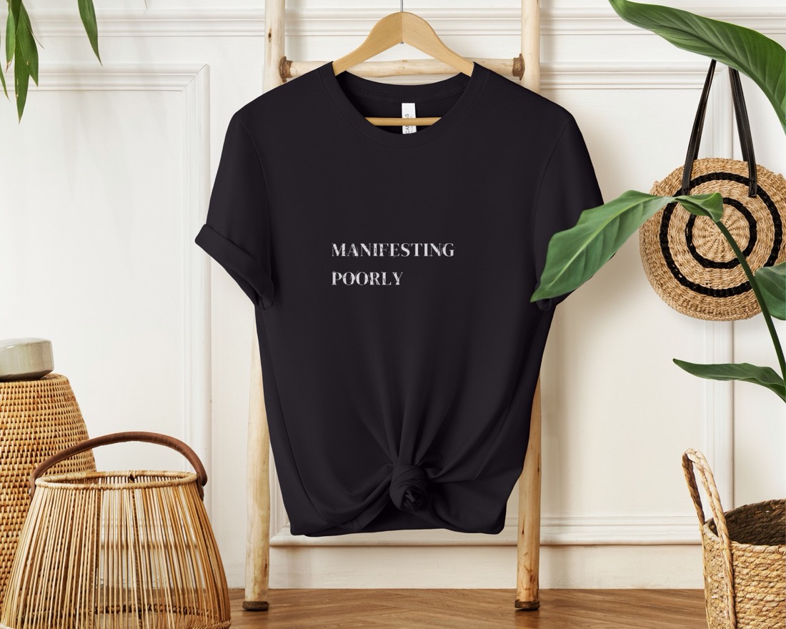 Black Manifesting Poorly tee hanging in a styled boho scene